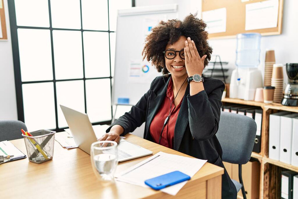 Afroamerykanka z afro włosami pracująca w biurze w słuchawkach operatora zakrywających jedno oko ręką, pewny siebie uśmiech na twarzy i niespodziewane emocje.  - Zdjęcie, obraz