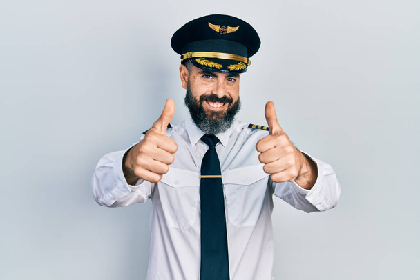 Fiatal spanyol férfi repülőgép pilóta egyenruhát visel, pozitív gesztust tesz kézzel, felemeli a hüvelykujját, mosolyog és örül a sikernek. győztes gesztus.  - Fotó, kép