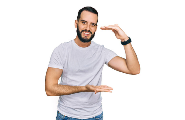Mladý muž s vousy na sobě ležérní bílé tričko gestikuluje s rukama ukazuje velké a velké velikosti znamení, opatření symbol. s úsměvem na kameru. koncepce měření.  - Fotografie, Obrázek