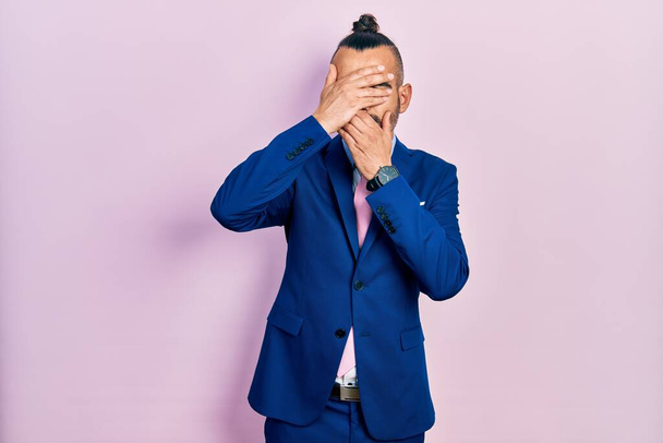 Young hispanic man wearing business suit and tie covering eyes and mouth with hands, surprised and shocked. hiding emotion  - 写真・画像