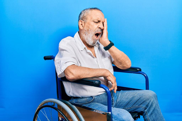 Bonito homem idoso com barba sentado em cadeira de rodas bocejando cansado cobrindo metade do rosto, olho e boca com a mão. cara dói na dor.  - Foto, Imagem