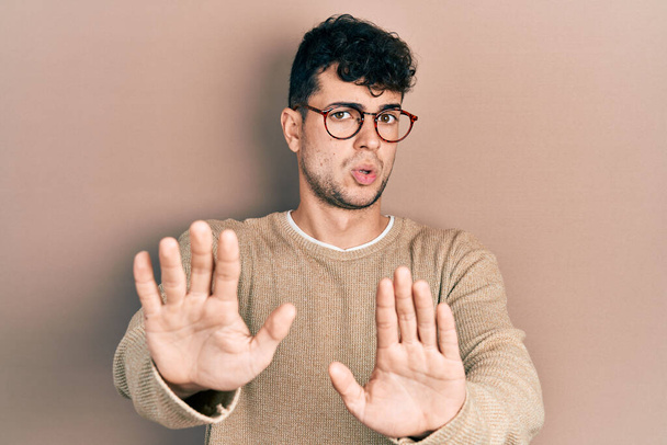 Jonge Spaanse man met casual kleding en een bril die handpalmen weghoudt en weigering en ontkenning toont met een angstige en walgelijke uitdrukking. stoppen en verbieden.  - Foto, afbeelding
