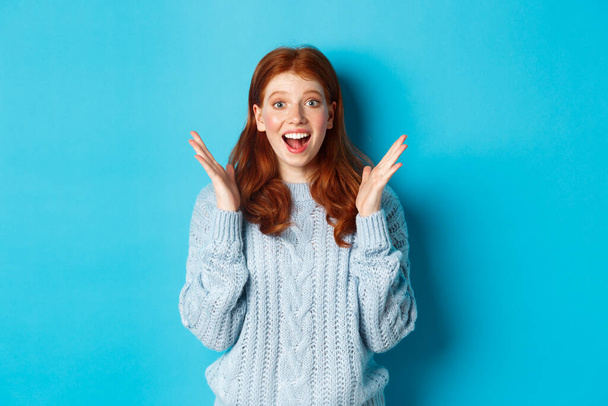 Surprised and happy redhead girl clap hands and staring at camera, smiling amazed, standing against blue background - Photo, Image