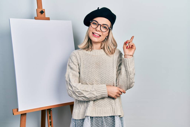 Piękna biała kobieta stoi przy sztaludze malarza uśmiechając się szczęśliwy wskazując ręką i palcem w bok  - Zdjęcie, obraz