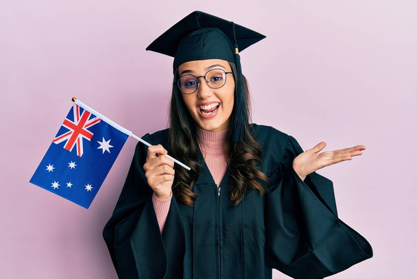 Young hispanic woman wearing graduation uniform holding australia flag celebrating achievement with happy smile and winner expression with raised hand  - Photo, Image