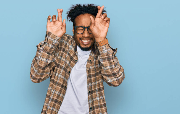 Νεαρός Αφροαμερικάνος με γενειάδα που φοράει καθημερινά ρούχα και γυαλιά να κάνει το δάχτυλο σταυρωμένο χαμογελώντας με την ελπίδα και τα μάτια κλειστά. τύχη και προληπτική έννοια.  - Φωτογραφία, εικόνα