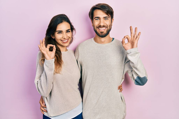 若いヒスパニック系のカップルは、顔に笑顔で自信を持って見えるカジュアルな服を着て,大丈夫なジェスチャーと幸せを示します.  - 写真・画像