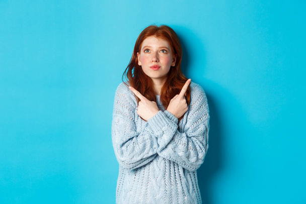 Excited redhead girl pointing fingers sideways, showing two choices and looking tempted up, standing against blue background - Photo, Image