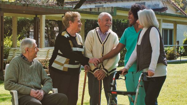 Enfermeira africana conversando com dois casais idosos aposentados ao ar livre no jardim do hospital de reabilitação. Concepção de felicidade, reabilitação e reforma. - Foto, Imagem