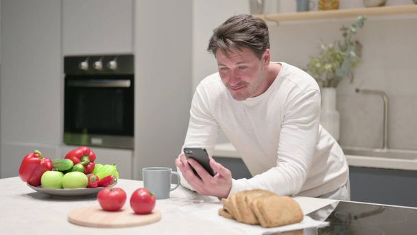 Μεσήλικας άνθρωπος που χρησιμοποιεί Smartphone, ενώ στην κουζίνα - Φωτογραφία, εικόνα