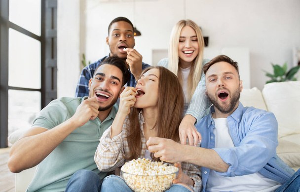 Разнообразные молодые друзья едят попкорн во время просмотра телевизора, веселятся вместе дома - Фото, изображение