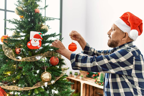 Senior man with beard wearing santa claus hat celebrating christmas at home, putting christmas decoration on christmas tree - Photo, Image