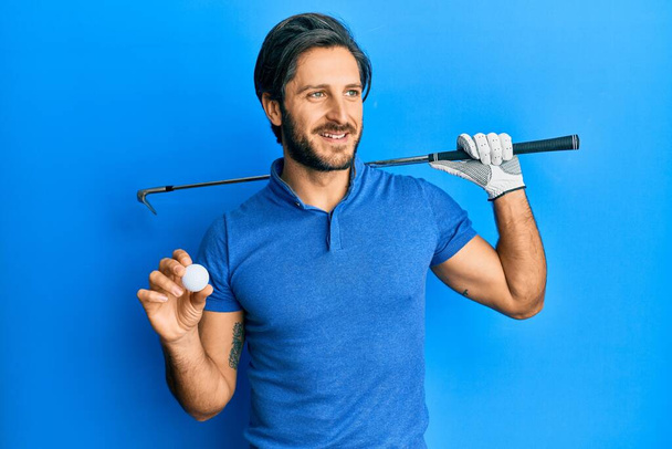 Joven hombre hispano sosteniendo pelota de golf sonriendo mirando hacia un lado y mirando hacia otro pensando.  - Foto, Imagen