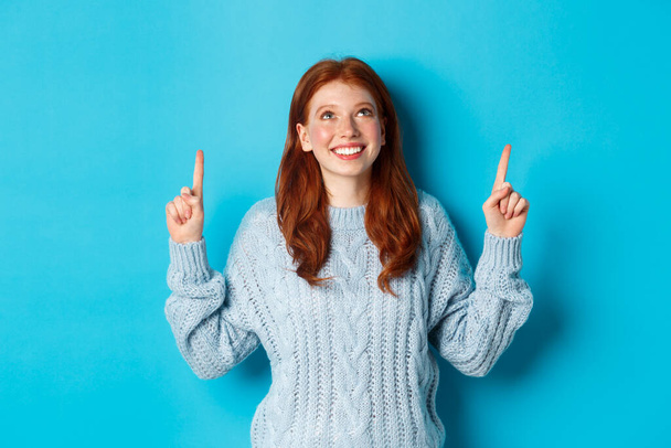 Winter holidays and people concept. Cute teenage redhead girl pointing fingers up, looking at top promo and smiling amused, standing over blue background - Fotoğraf, Görsel