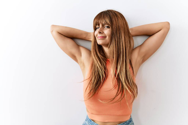 Латиноамериканка с причёской, стоящая на изолированном фоне расслабляющая и растягивающая, руки и руки за головой и шеей улыбаются счастливо  - Фото, изображение
