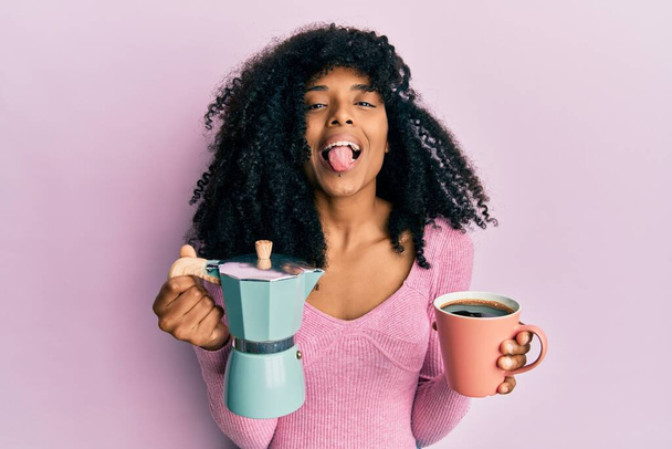 Mujer afroamericana con cabello afro bebiendo café italiano sacando la lengua feliz con expresión divertida.  - Foto, Imagen