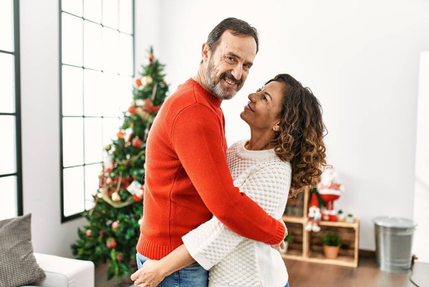 Casal hispânico de meia idade sorrindo feliz e abraçando celebrando o Natal em casa. - Foto, Imagem
