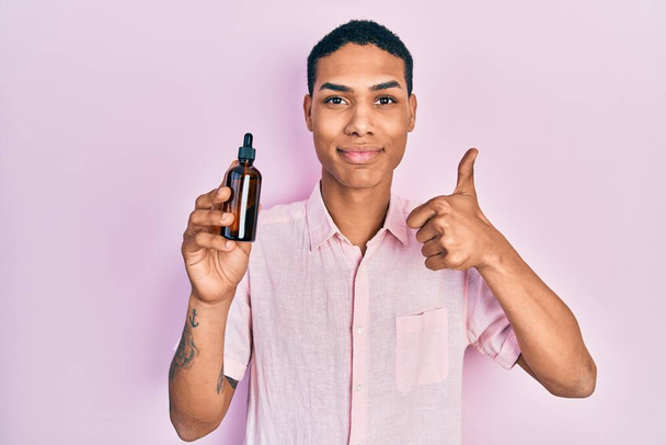 Young african american guy holding serum bottle smiling happy and positive, thumb up doing excellent and approval sign  - Fotó, kép