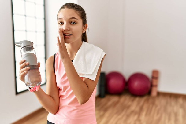 Młoda brunetka nastolatka w odzieży sportowej trzymająca butelkę wody za usta, mówiąca pogłoski, szepcząca złośliwe rozmowy.  - Zdjęcie, obraz