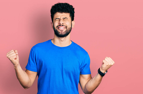 Young arab man with beard wearing casual blue t shirt very happy and excited doing winner gesture with arms raised, smiling and screaming for success. celebration concept.  - Fotó, kép