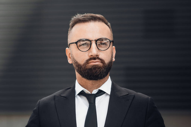 Business lifestyle concept. Portrait of bearded mature businessman in glasses and black suit posing to camera outdoors - Fotó, kép