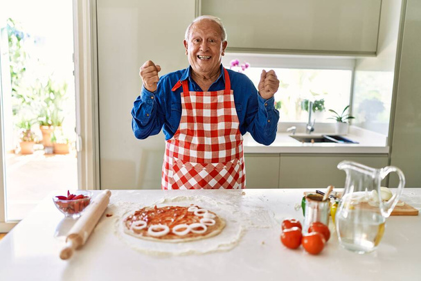 Senior άνθρωπος με γκρίζα μαλλιά μαγείρεμα πίτσα στο σπίτι κουζίνα ουρλιάζοντας υπερήφανος, γιορτάζει τη νίκη και την επιτυχία πολύ ενθουσιασμένος με σήκωσε τα χέρια  - Φωτογραφία, εικόνα