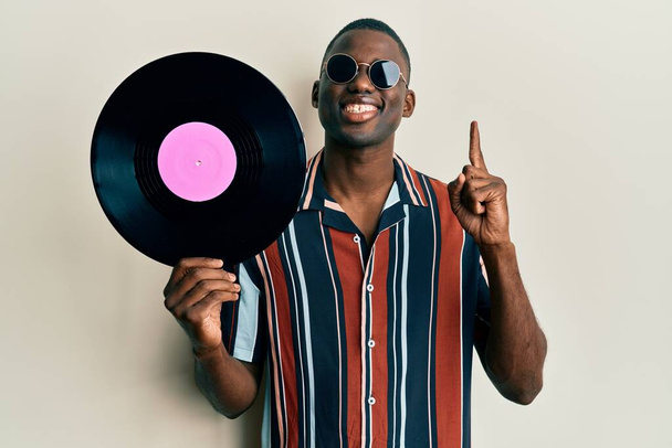 Young african american man wearing hippie style holding vinyl disc smiling with an idea or question pointing finger with happy face, number one  - 写真・画像