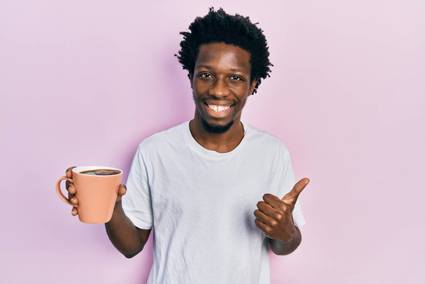 Młody Afroamerykanin trzyma filiżankę kawy uśmiechnięty szczęśliwy i pozytywny, kciuk do góry robi doskonałe i znak aprobaty  - Zdjęcie, obraz