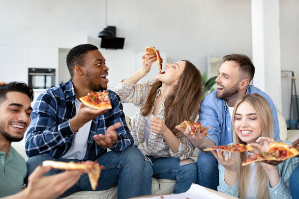 Cool diverse young friends eating pizza together, laughing and chatting at home - Foto, afbeelding