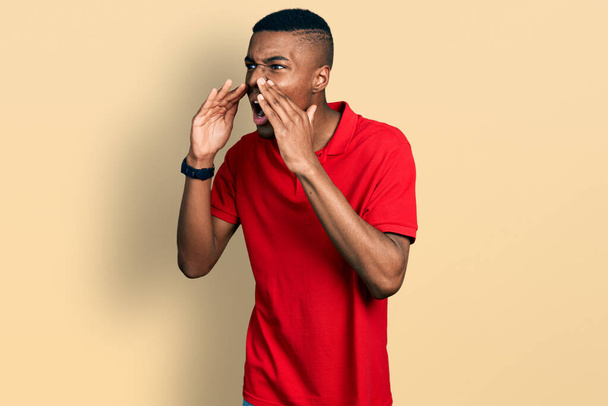 Молодой африканский американец в обычной красной футболке кричит сердито с руками над ртом  - Фото, изображение