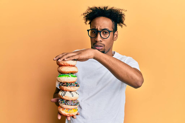 Young african american man with beard holding pile of tasty colorful doughnuts clueless and confused expression. doubt concept.  - Фото, изображение