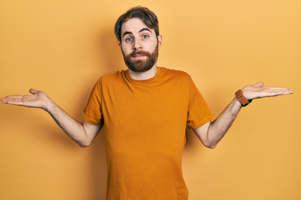 Hombre caucásico con barba con camiseta amarilla casual expresión despistada y confusa con los brazos y las manos levantadas. concepto de duda.  - Foto, Imagen