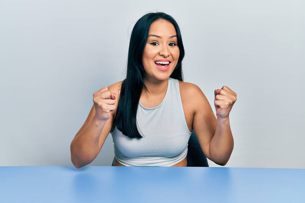 Beautiful hispanic woman with nose piercing sitting on the table celebrating surprised and amazed for success with arms raised and open eyes. winner concept.  - Photo, image