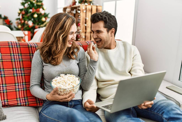 Young hispanic couple watching film eating popcorn sitting on the sofa at home. - Photo, Image