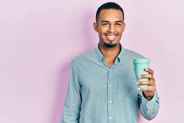 Νεαρός Αφροαμερικάνος που πίνει ένα φλιτζάνι καφέ και δείχνει θετικός και χαρούμενος που στέκεται και χαμογελάει με αυτοπεποίθηση που δείχνει τα δόντια του  - Φωτογραφία, εικόνα