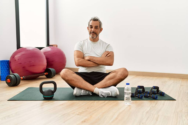 Hombre hispano de mediana edad sentado en la alfombra de entrenamiento en el gimnasio cara feliz sonriendo con los brazos cruzados mirando a la cámara. persona positiva.  - Foto, Imagen
