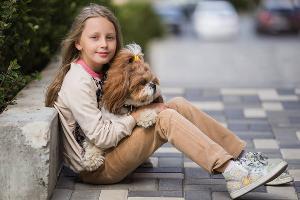 Portrait of beautiful preteen girl petting and hugging shih tzu dog looking at the camera outdoors. - Fotó, kép