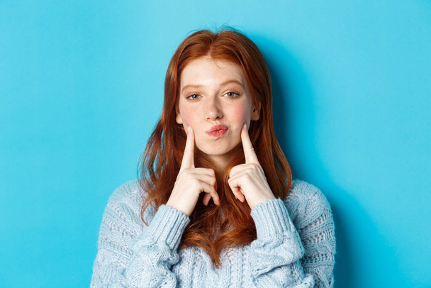 Nahaufnahme des niedlichen rothaarigen Mädchens im Pullover, aufgerissene Lippen und Zeigefinger auf Wangen, stochernde Grübchen, vor blauem Hintergrund stehend - Foto, Bild