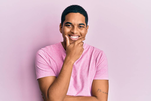 Young handsome hispanic man wearing casual pink t shirt looking confident at the camera smiling with crossed arms and hand raised on chin. thinking positive.  - Valokuva, kuva