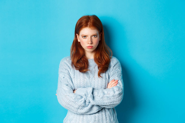 Image of angry redhead girl feeling offended, cross arms on chest and sulking, staring at camera mad, standing against blue background - Fotoğraf, Görsel