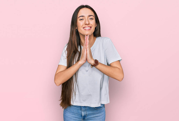 Nuori latino tyttö yllään rento valkoinen t paita rukoilee kädet yhdessä pyytäen anteeksiantoa hymyillen luottavainen.  - Valokuva, kuva