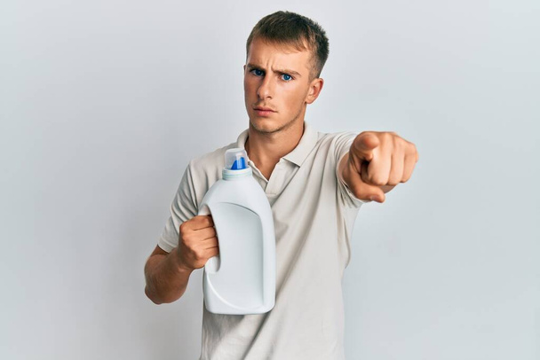 Junger Kaukasier mit Waschmittelflasche, die mit dem Finger in die Kamera und zu dir zeigt, selbstbewusste Geste, die ernst wirkt  - Foto, Bild