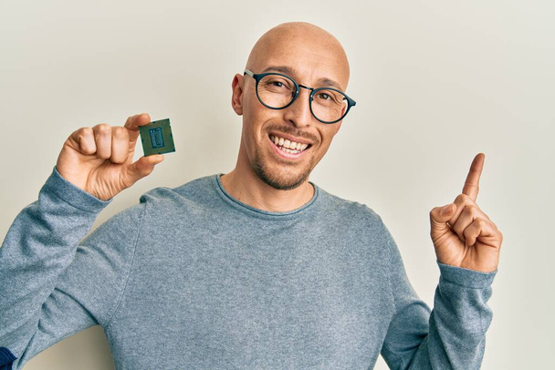 Bald man with beard holding cpu computer processor smiling happy pointing with hand and finger to the side  - Foto, afbeelding
