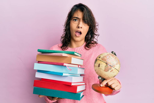 Jonge Spaanse vrouw met een stapel boeken en wereldbal in shock gezicht, op zoek sceptisch en sarcastisch, verrast met open mond  - Foto, afbeelding