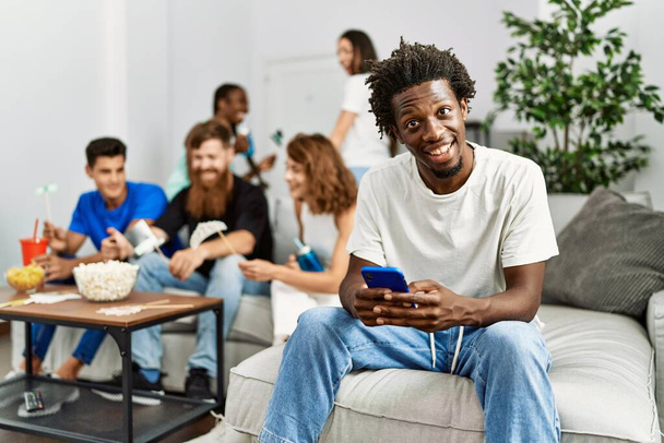 Eine Gruppe junger Freunde sitzt glücklich lächelnd auf dem Sofa. Frau nutzt Smartphone zu Hause. - Foto, Bild