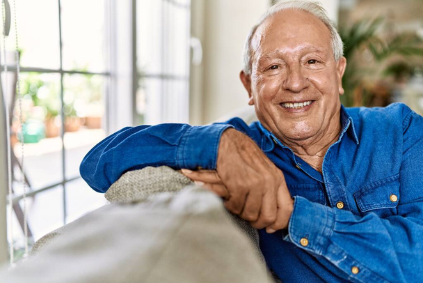 Starszy mężczyzna z siwymi włosami siedzący na kanapie w salonie swojego domu. Dojrzały mężczyzna uśmiechnięty szczęśliwy w domu. - Zdjęcie, obraz