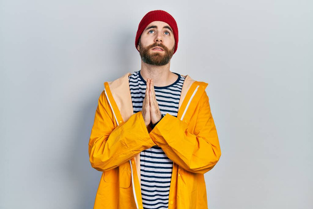 Homem caucasiano com barba vestindo capa de chuva amarela implorando e orando com as mãos junto com expressão de esperança no rosto muito emocional e preocupado. implorando.  - Foto, Imagem