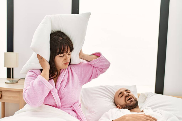 Hispánka naštvaná s polštářem na uších, zatímco muž chrápe na posteli. - Fotografie, Obrázek