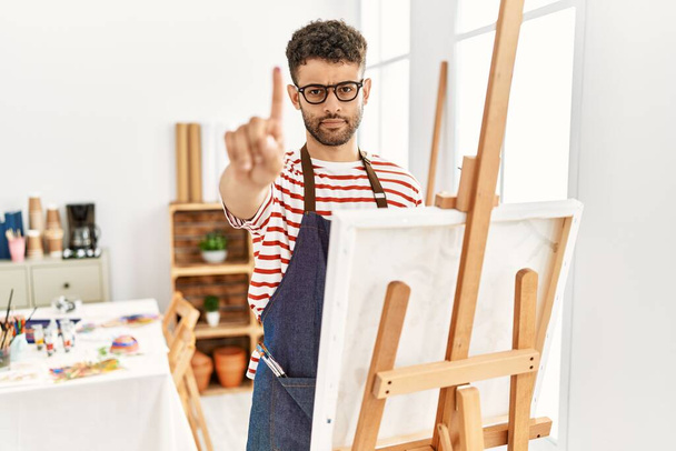 Арабский молодой человек в художественной студии показывает пальцем вверх и сердитым выражением лица, не показывая жеста  - Фото, изображение