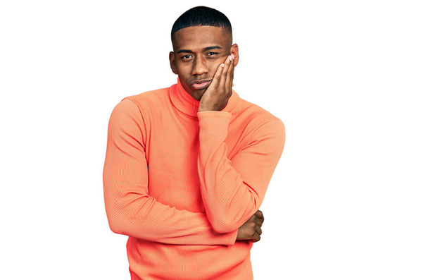 Jeune homme noir portant un pull à col roulé orange pensant fatigué et ennuyé par des problèmes de dépression avec les bras croisés.  - Photo, image